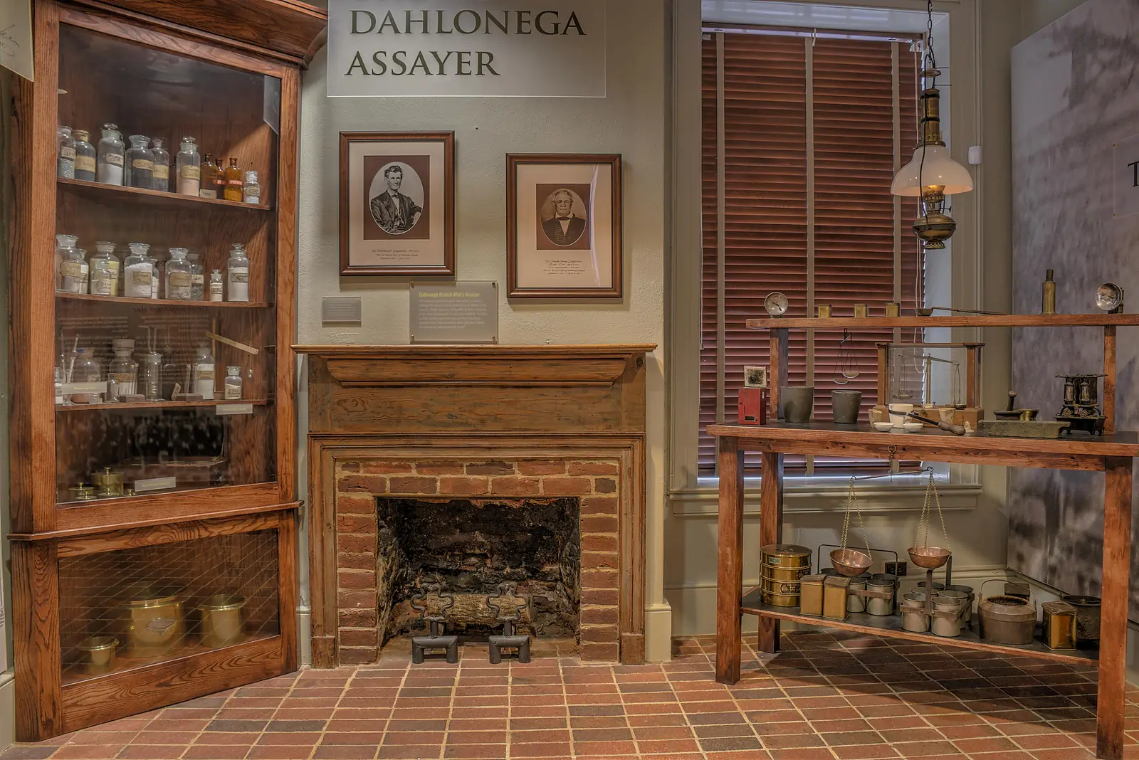 A museum display headed 'Dahlonega Assayer': a cabinet of glass chemical bottles, two framed portraits over a brick fireplace, and a table with balance scales, crucibles and brass tins.