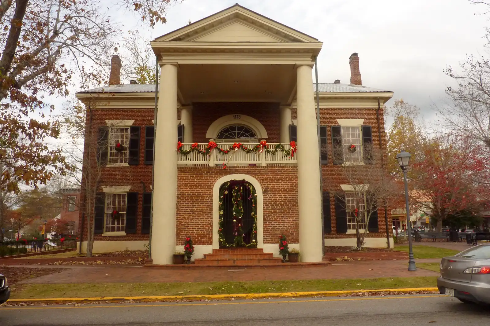 Front view of the courthouse with its four white columns and balcony, flanked by trees in russet and red autumn leaf under a grey sky. A Christmas garland frames the door and balcony; part of a parked car is at the right edge.