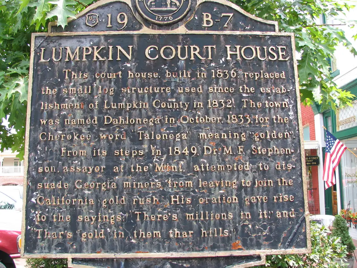 Close view of the weathered black and gold state historical marker 'Lumpkin Court House', whose text gives the 1836 construction date, the naming of Dahlonega and the origin of 'Thar's gold in them thar hills'. A US flag and shopfront are behind.