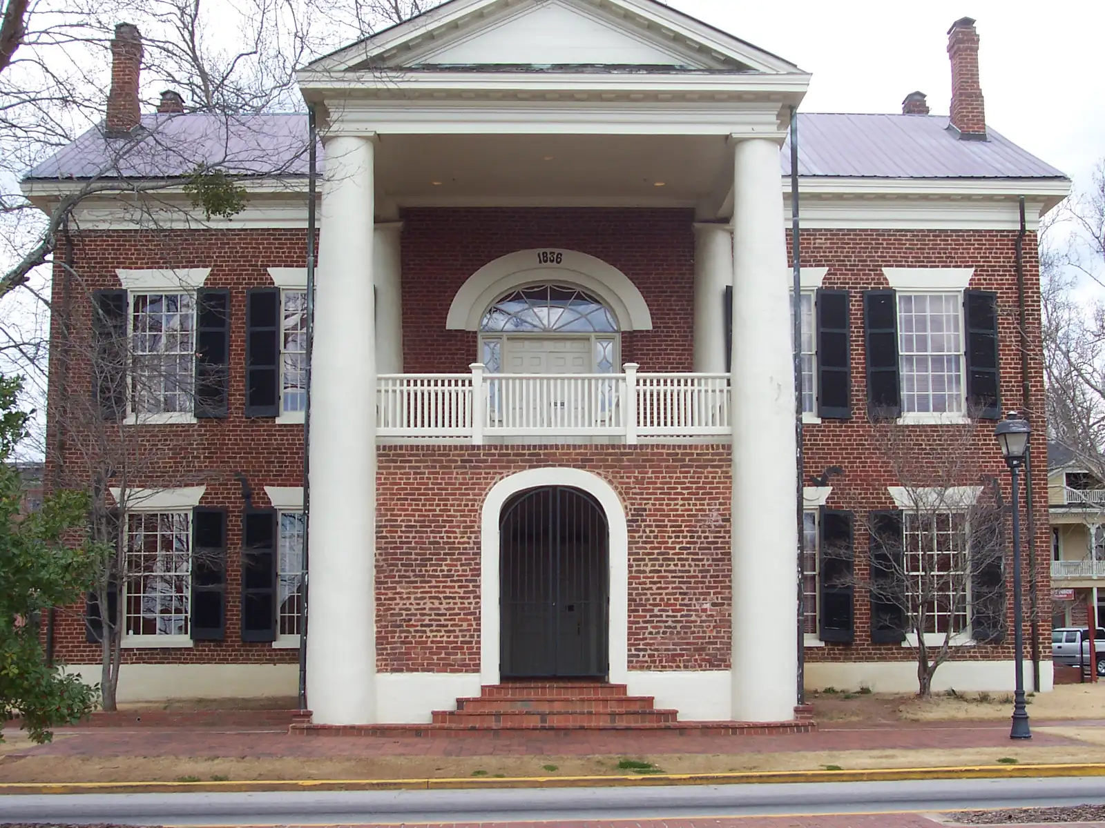 Straight-on view of the two-storey red brick courthouse front: four white columns, white balcony, arched iron gate and the date 1836 over the fanlight. Bare trees and an overcast sky.