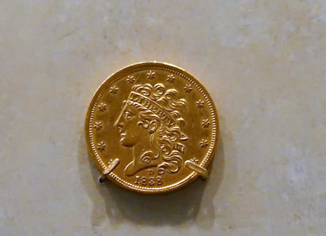 A gold five-dollar coin with a Liberty head, thirteen stars, a small 'D' mint mark and the date 1838, held by two clips on pale stone above engraved wall lettering that reads 'United States 5 Dollars 1838 D'.