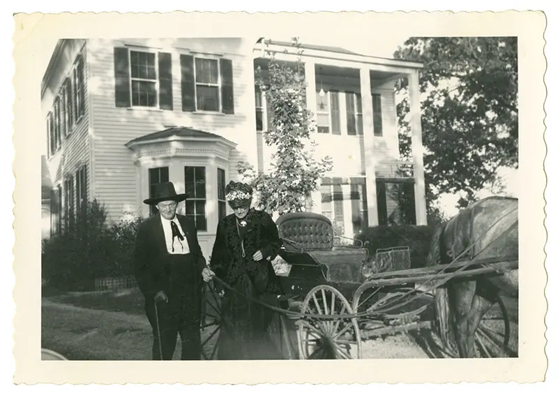 Black and white snapshot with a deckled border: an elderly man in a dark suit and wide hat and a woman in a dark period dress and bonnet stand beside a horse-drawn buggy in front of a white two-storey house.