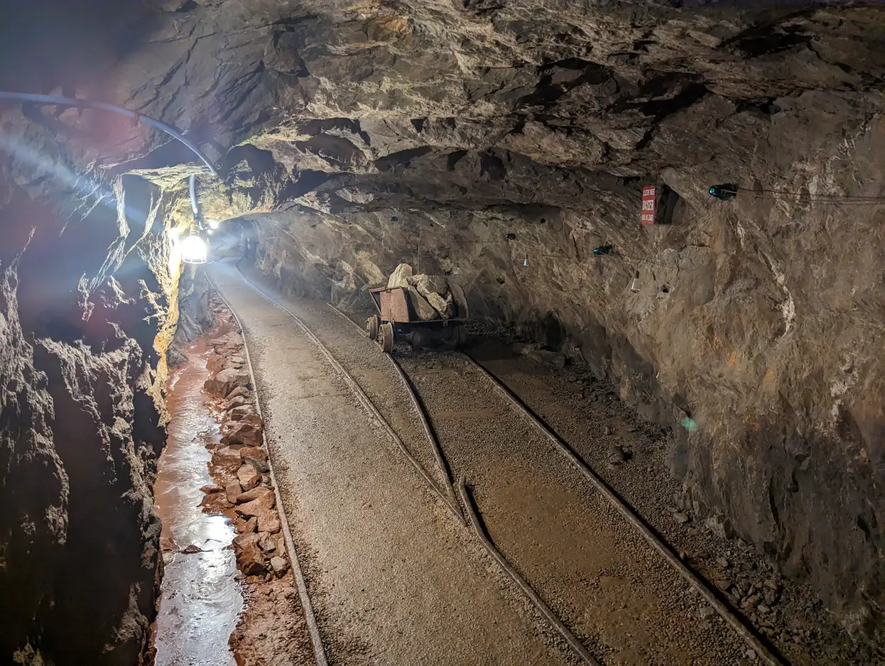 Inside a rock-walled mine tunnel lit by bare bulbs: narrow-gauge rails curve away past a small ore cart, with a rust-coloured drainage channel along the left wall.