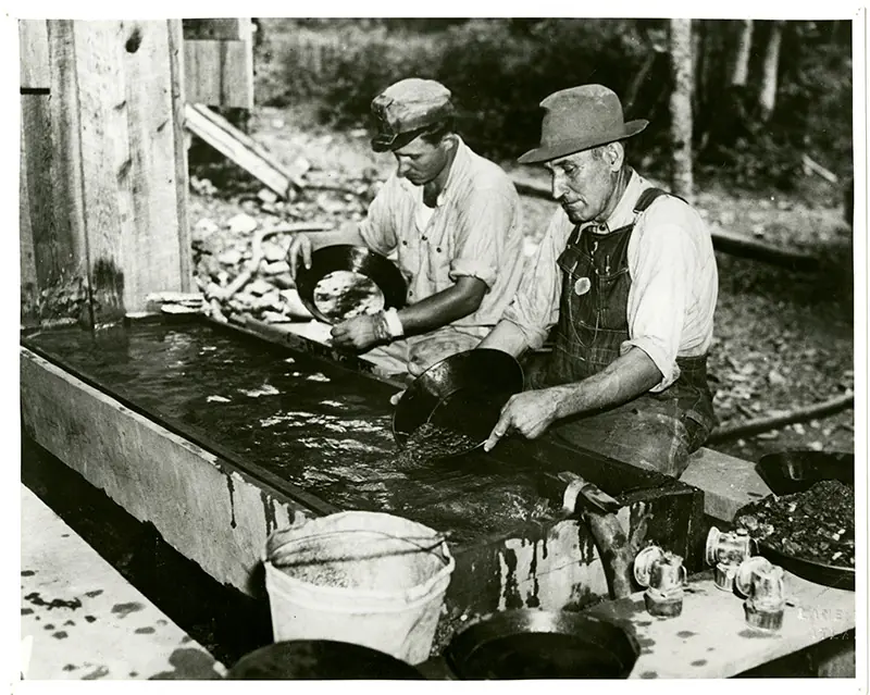 Two men in work clothes, one in a brimmed hat and overalls, sit at a long wooden water trough swirling gold pans; more pans, a bucket and glass jars are in the foreground.