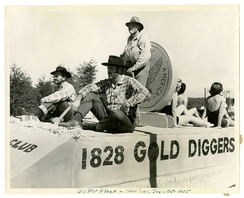Black and white close view of a parade float lettered '1828 GOLD DIGGERS': three men in hats and work shirts with a pick, a giant coin prop, and two women in swimsuits seated behind. A handwritten caption on the print border reads 'B+PW Float - Gold Rush Day - Oct 1955'.