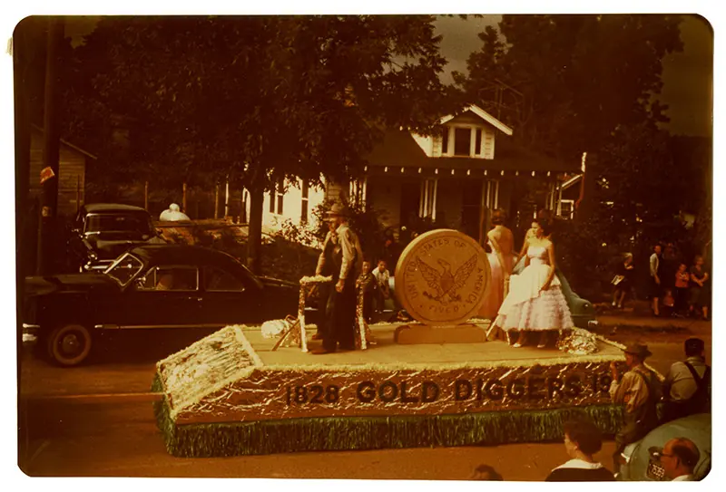 A faded colour snapshot of a parade float lettered '1828 GOLD DIGGERS' carrying a giant gold coin, a man in prospector dress and a woman in a white ball gown, passing 1950s cars, a bungalow and spectators.