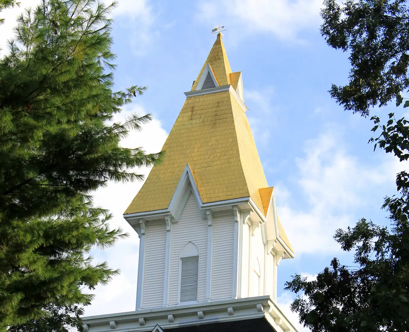 Portrait-format close view of the gold-leafed steeple and white belfry above the brick gable and columned porch, with US and Georgia flags, framed by green trees against a blue sky with clouds.