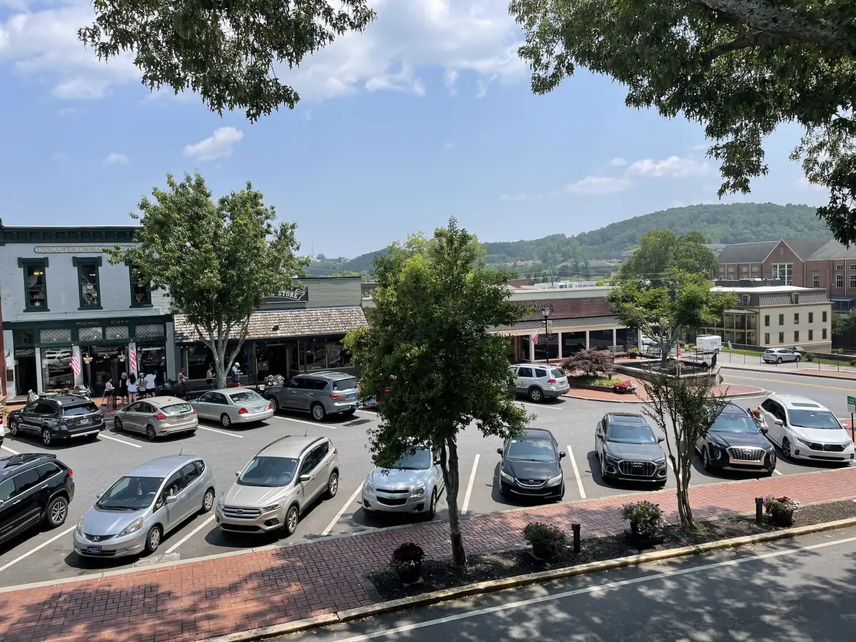 Elevated summer view across the brick-edged public square: angled parking full of cars, two-storey storefronts including a painted brick building with green trim, street trees, and wooded hills behind. No crowd and no event.