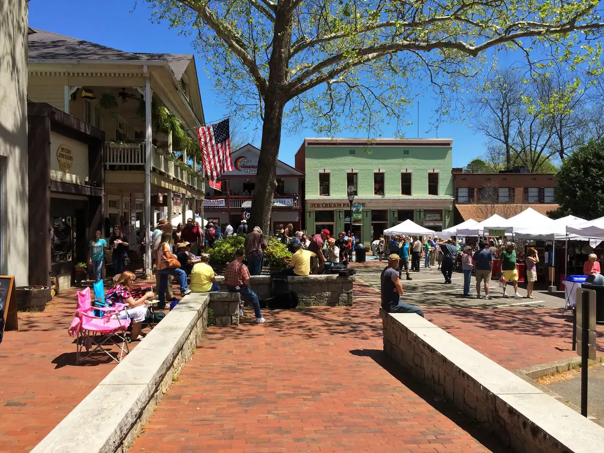 A busy spring day on the brick-paved square: a crowd around musicians seated on a stone wall, white vendor tents in the street, a US flag on a balconied building, and a pale green two-storey storefront with an ice cream parlour sign under a blue sky.